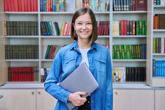 Portrait of smiling young female university student looking at camera, standing posing with laptop in library classroom of educational building. Knowledge, higher education, youth concept