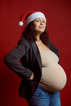 Studio portrait of emotional charismatic curly haired pregnant woman in stylish blazer, blue jeans and Santa hat, smiling cutely looking at camera, isolated red color backdrop. Happy New Year concept