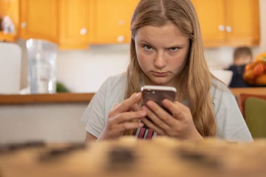 A teenage girl of European appearance, sits in the living room at a table in her hands, holds a phone, looks surprised at the camera. Portrait of a girl with blond hair in the house.High quality photo