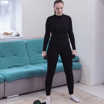 girl at home and exercising with dumbbells. The concept of sports and recreation. A beautiful woman in sportswear with black dumbbells in her hands. In the background is a sofa and a sleeping cat