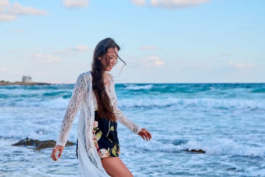 Beautiful young woman in lace dress walking along beach, copy space. Smiling Asian lady enjoying sea nature, summer vacation. Travel, tourism, holiday, beauty, naturalness, fashion, people concept