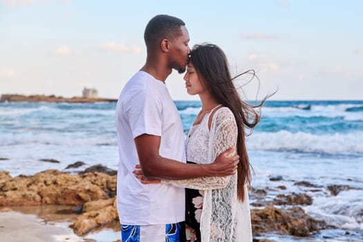 Kiss of loving young couple, sea sky background. Beautiful multicultural couple, profile view, honeymoon at seaside resort, vacation together. Love, relationship, tourism travel, nature beauty people