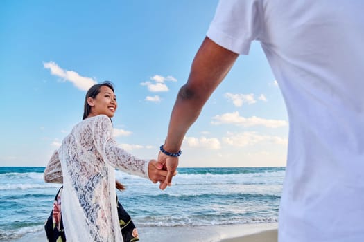 Loving happy couple walking holding hands on beach. Asian woman holding man's hand, sea sky background. Love, relationship, vacation, tourism, tourist travel to seaside resort, honeymoon concept