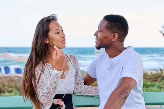 Young multicultural couple smiling talking sitting on bench, sea background. Beautiful asian woman and african american man together outdoor, sea vacation together, love, relationship, communication