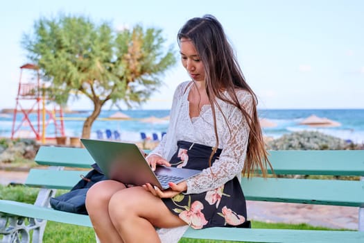 Beautiful young Asian woman using laptop sitting on an outdoor bench. Focused serious female looking in laptop screen, exotic nature sea background. Freelance, remote work study, technology, lifestyle