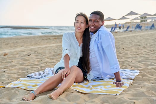 Happy multiethnic family resting on beach together. Asian woman and African man seated hugging each other on sand. Relationships, vacation travel and tourism at seaside resorts, people 20 30 years old