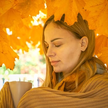a girl of European appearance with blond hair, stands in brown in autumn under a tree with brown leaves with a cup in her hands. Autumn photo session. High quality photo