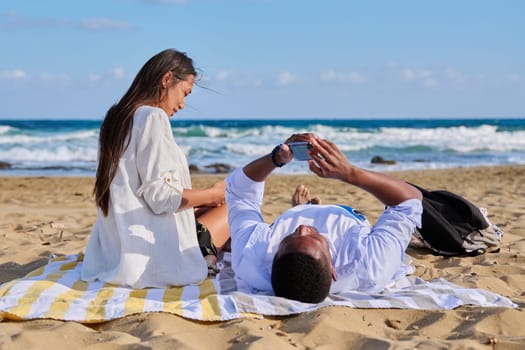 Young multicultural couple lying on beach using smartphones. Relaxing tourists lying on beach towels looking at smartphones. Lifestyle, technology, tourism, travel, nature, leisure, people concept