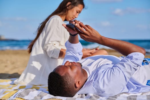 Young multicultural couple lying on beach using smartphones. Relaxing tourists lying on beach towels looking at smartphones. Lifestyle, technology, tourism, travel, nature, leisure, people concept