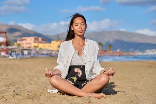 Young woman in lotus position on the beach practicing yoga. Natural Asian female meditating with closed eyes, sitting on the sand. Healthy body, harmony, beauty, peace of mind, mental health concept