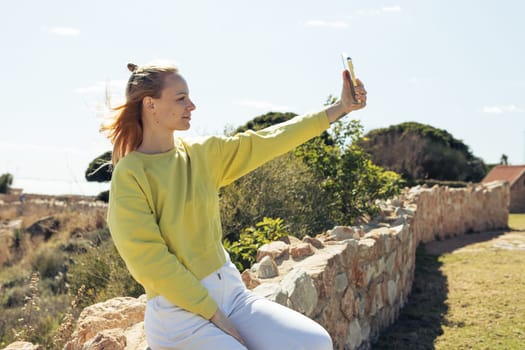 a beautiful girl sits on a fence with stones and takes a selfie against the blue sky .beautiful landscape with a girl. High quality photo