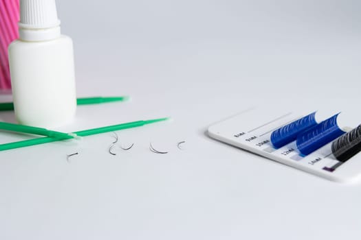 Artificial eyelashes in black and blue are pasted on a white tablet on a white background. On the left side are bundles of eyelashes, pink and green microbrushes and a white bottle of liquid.close-up