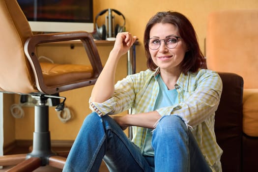 Middle aged woman freelancer, workplace in home office. Female resting, sitting on floor near table with computer and armchair. Lifestyle, home, interior, people 40s age concept