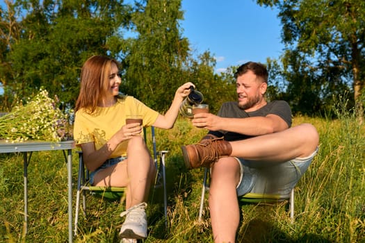 Happy middle-aged married couple resting outdoors, in meadow nature. Man and woman sitting on folding camping chair drinking coffee enjoying wildlife scenery sunbathing talking together