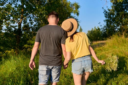 Happy romantic couple, man and woman walking together holding hands, female with bouquet of wildflowers, natural landscape in sunset, back view. Relationships, love, dating, people 30s 40s age