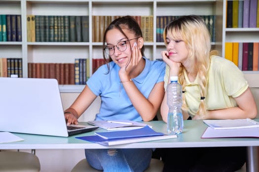 Two teenage schoolgirls students sitting with a laptop in the library, girls looking at laptop screen. Education, high school, technology, teamwork, learning, e-learning, adolescence concept