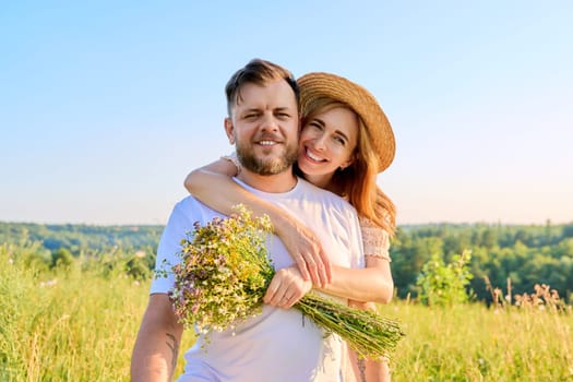 Happy middle-aged couple in love embracing, with bouquet of wildflowers, outdoors on summer nature meadow, looking at camera. Date, anniversary, family, wedding, celebration, relationship, 40s people