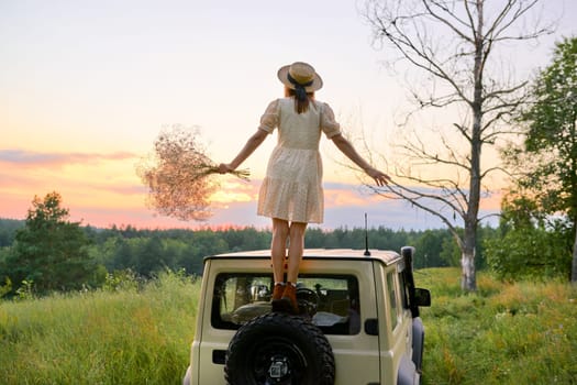 Summer, beauty of nature, adventure, travel, trip, freedom, journey. Beautiful woman in straw hat, dress enjoying setting sun, landscape, pink sky, on SUV car, wildlife background back view