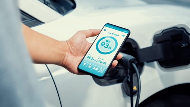 Businessman holding smartphone display battery status interface by smart EV mobile application while EV car recharging electricity from charging station in car park. Peruse