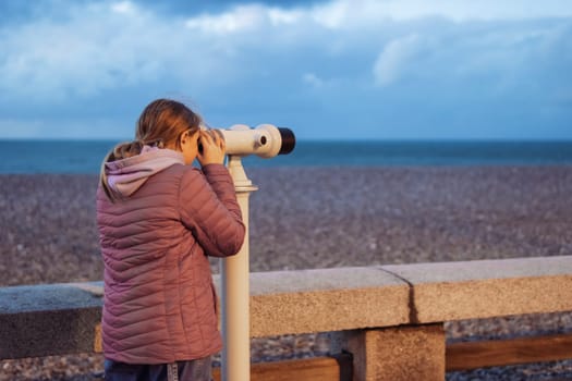 a girl of European appearance with blond hair stands on the embankment looking through binoculars at the ocean. the girl is dressed in a pink jacket and jeans, there is a place for. High quality photo