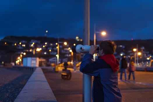 a boy of European appearance with blond hair stands on the embankment in the evening looks through binoculars at the ocean. the boy is dressed in a jacket there is a place for. High quality photo