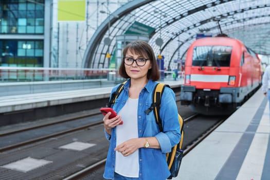 Woman waiting for railway public electric transport on platform of city station. Female with smartphone using banking app to buy online ticket payment, online timetable and route service, technology