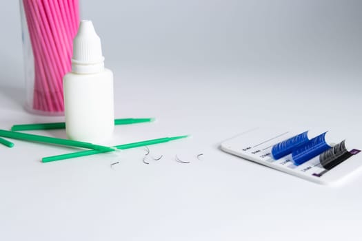 Artificial eyelashes in black and blue are pasted on a white tablet on a white background. On the left side are bundles of eyelashes, pink and green microbrushes and a white bottle of liquid