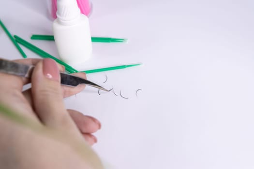 A woman's hand holds an eyelash extension tweezers and takes artificial eyelashes. Eyelash extension materials lie nearby