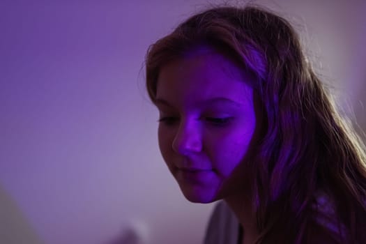 teenage girl of European appearance with blond hair, sits in a dark room with a colored light turned on, a purple light falls on the girl's face, there is a place for an inscription.High quality photo