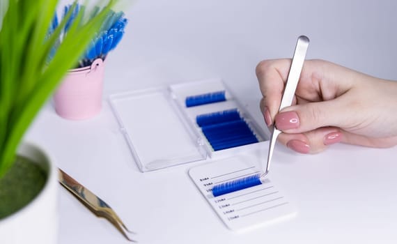 Materials for eyelash extensions.a girl holds tweezers and blue eyelashes lie on a table on a white background.nearby lies gold-colored tweezers and in the background a pink bucket with brushes