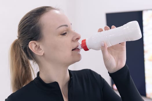 Portrait close-up of a young tired caucasian fitness woman standing in black sportswear and drinking water from a white bottle isolated on white background. Home workout concept