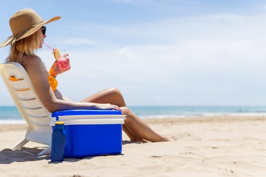 Happy young european girl with a cocktail in her hands is resting in a sun lounger on the sea near the blue refrigerator, there is space for an inscription. High quality photo