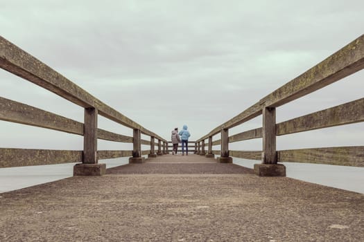 two girls stand in jackets on a bridge in the distance and look at the ocean, stand with their backs to the camera, there is a place for an inscription. High quality photo