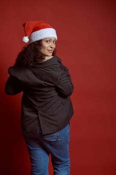 Back view of a beautiful multi ethnic young adult pregnant woman, wearing Santa hat, blue jeans and gray blazer, smiles cutely looking at camera, isolated over red color studio background