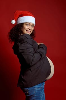 Authentic portrait curly haired pretty woman, pregnant expectant mother in Santa hat, smiles looking at camera, expressing positive emotions, isolated on red studio background. Happy New Year concept