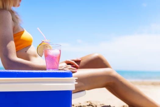 A tanned thin girl in a bikini sits on a sun lounger close-up on slender legs, there is a portable refrigerator, there is a glass with a cocktail on it, a beautiful beach landscape,High quality photo