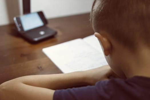 a boy with a European appearance with blond hair is focused on doing homework in a children's room on distance learning in front of the phone and writing in a notebook with a pen