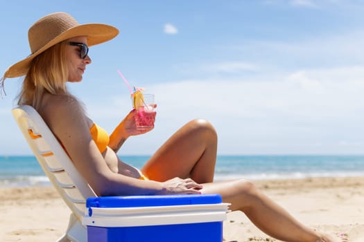 Happy young european girl drinks a cocktail and rests in a sun lounger on the beach, space for an inscription portable blue refrigerator by the sea. High quality photo