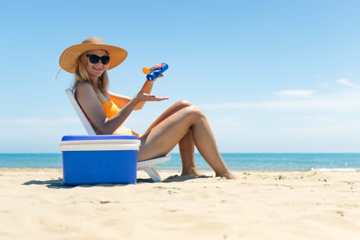 a tanned girl in a swimsuit sits on a sun lounger smears sunblock on her legs side view on the beach and there is a blue portable refrigerator for drinks nearby. Summer vacation High quality photo
