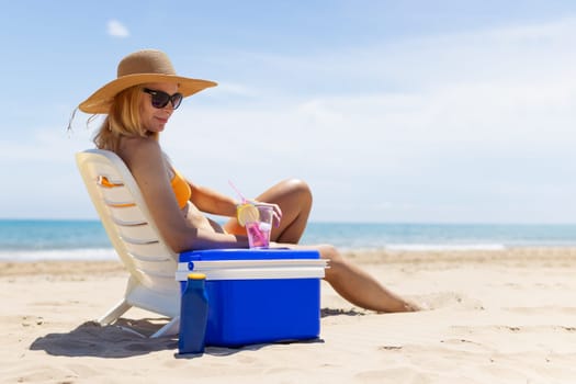 Happy young european girl drinks a cocktail and rests in a sun lounger on the beach, space for an inscription portable blue refrigerator by the sea. High quality photo