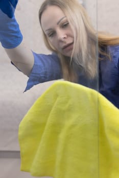 blond girl with European appearance washing glass with yellow cloth in blue gloves at home.House cleaning concept