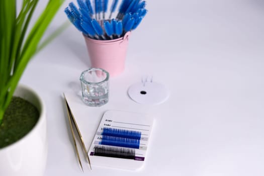 Materials for eyelash extensions. Tweezers, artificial blue and black eyelashes. Nearby is a paint machine and blue brushes in a pink bucket. On the side there is a place for an inscription