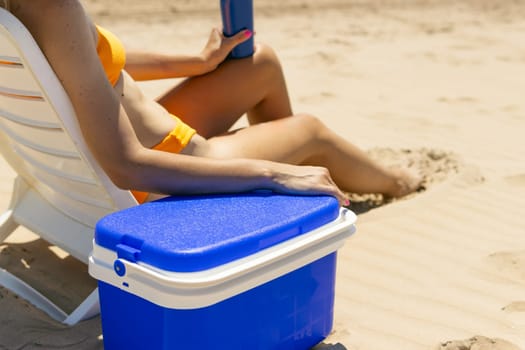 beach background with girl,slender girl in a swimsuit sits on a sun lounger smears her legs with sunblock, a blue portable refrigerator for drinks stands nearby. High quality photo.