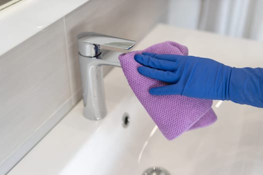 Woman cleaning the faucet with purple sponge in the bathroom wearing blue gloves white sink close up. House cleaning concept