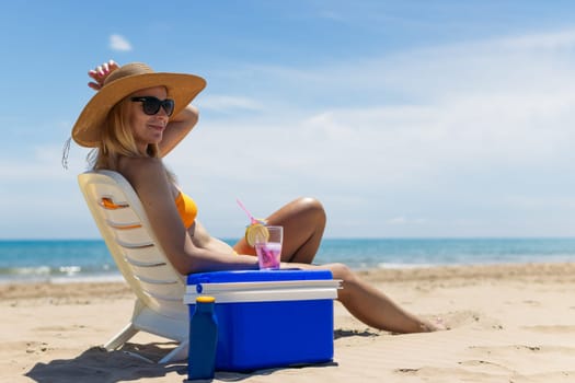 a slender blonde in a bright bikini, sunglasses and a hat is sunbathing on a sun lounger with a pink cocktail in her hand by the sea, next to a portable drinks fridge. . High quality photo