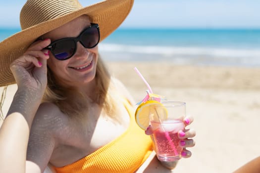 A sexy blonde in a bright bikini is sunbathing on a sun lounger with a cocktail in her hand by the sea. A beautiful girl is enjoying her summer holidays at an expensive resort, High quality photo