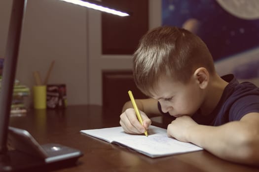 a boy with a European appearance with blond hair is focused on doing homework in a children's room on distance learning in front of the phone and writing in a notebook with a pen