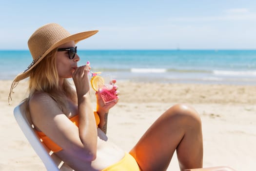 Happy young european girl drinks a cocktail and rests in a sun lounger on the beach, space for an inscription by the sea. High quality photo