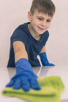 a little boy of European appearance with blond hair does house cleaning in blue gloves and a yellow napkin washes a glass white glossy table in the living room