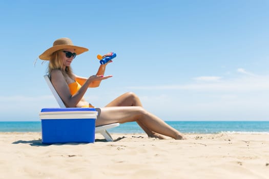 beach background with a girl, a slender girl in a swimsuit sits on a sun lounger smears her legs with sunblock, a blue portable refrigerator for drinks stands nearby. High quality photo